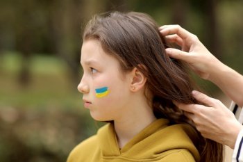 Close up of caring young mother braids hair sad little daughter with flag on face outdoors. Peace, no war, help, stop russian aggression. Invasion of Russia in Ukraine. High quality photo. Close up of caring young mother braids hair sad little daughter with flag on face outdoors. Peace, no war, help, stop russian aggression. Invasion of Russia in Ukraine. High quality photo.