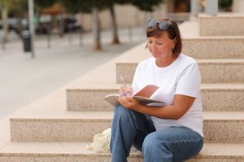 mature woman works remotely, higher education for adults, casually female writes and makes note in a notebook sitting on stairs outdoors, freelancer.College study program, academic educational course. mature woman works remotely, higher education for adults, casually female writes and makes note in a notebook sitting on stairs outdoors, freelancer. College study program, academic educational course