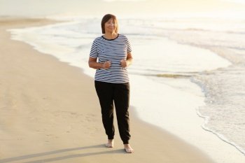 Smiling middle aged woman running on the beach on sunrise. 40s or 50s attractive mature lady in sports clothes doing jogging workout enjoying fitness and healthy lifestyle at beautiful sea landscape. Smiling middle aged woman running on the beach on sunrise. 40s or 50s attractive mature lady in sports clothes doing jogging workout enjoying fitness and healthy lifestyle at beautiful sea landscape.