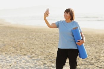 Smiling adult woman in training wear holding yoga mat make photo or have video call with smartphone over sea nature background. Middle aged blogger. Healthy lifestyle. Meditation, yoga and relaxation. Smiling adult woman in training wear holding yoga mat make photo or have video call with smartphone over sea nature background. Middle aged blogger. Healthy lifestyle. Meditation, yoga and relaxation.