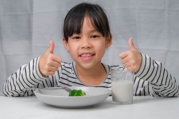 Cute Asian girl drinking a glass of milk in the morning before going to school. Little girl eats healthy vegetables and milk for her meals. Healthy food in childhood.