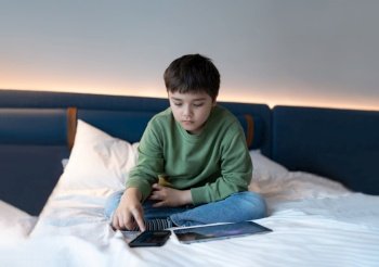 Education concept,Kid using mobile phone playing game on internet, Homeschooling Child doing homework online by tablet pad at home,Boy sitting on bed relaxing, watching cartoon or talking with friend