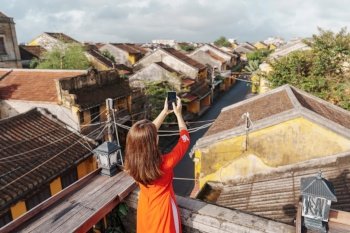 happy woman wearing Ao Dai Vietnamese dress, traveler taking photo by mobile phone on rooftop at Hoi An ancient town in Vietnam. Vietnam and Southeast travel concept
