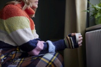 Woman In Gloves With Hot Drink Trying To Keep Warm By Radiator During Cost Of Living Energy Crisis