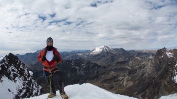Young male climber standing on the summit of snowy mountain wearing helmet, headlamp, red coat and rope during a cloudy day. Young male climber standing on the summit of snowy mountain wearing helmet, headlamp, red coat and rope