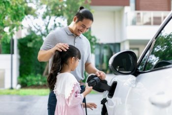 Progressive father and daughter returned from school in electric vehicle that is being charged at home. Electric vehicle driven by renewable clean energy. Home charging station concept for environment. Progressive concept of father and daughter with EV car and home charging station