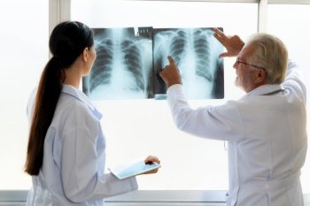 In a hospital sterile room, two professional radiographers hold and examine a radiograph for medical xray diagnosis. Novice doctor seeks advice on a patient’s condition from experienced older doctor.. Two doctors examine radiograph for medical xray diagnosis in sterile room.