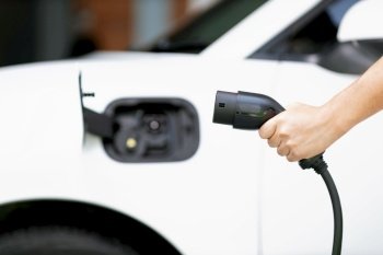 Focus hand holding EV charger plug with blurred background of progressive electric vehicle and socket parking in home garage with electric charging station powered by clean and sustainable energy.. Focus hand hold EV plug with blur image of progressive car at charging station.