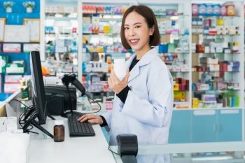 Portrait of young affable pharmacist and qualified pharmaceutical, medicine pill container or bottle mockup for copyspace at pharmacy. Druggist working with her diary job at drugstore, medicine shelf.. Portrait of young asian pharmacist pill bottle mockup at qualified pharmacy