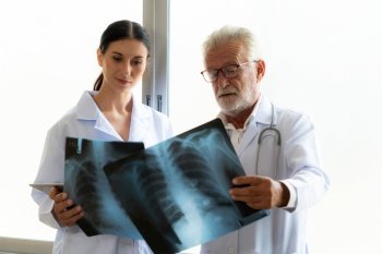 In a hospital sterile room, two professional radiographers hold and examine a radiograph for medical xray diagnosis. Novice doctor seeks advice on a patient’s condition from experienced older doctor.. Two doctors examine radiograph for medical xray diagnosis in sterile room.