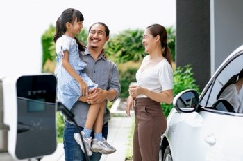 Progressive young parent teach daughter how to recharge or refuel EV car at home charging station. Green and clean energy from electric vehicle for healthy environment. Eco power from renewable source. Progressive young parents and daughter living in a home with an electric car.