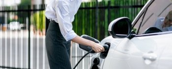 Closeup progressive suit-clad businesswoman with her electric vehicle recharge her car on public charging station in modern city with power cable plug and renewable energy-powered electric vehicle.. Closeup progressive businesswoman with electric vehicle at charging station.