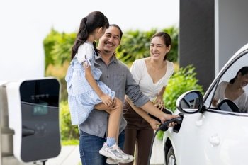 Progressive young parent teach daughter how to recharge or refuel EV car at home charging station. Green and clean energy from electric vehicle for healthy environment. Eco power from renewable source. Progressive young parents and daughter living in a home with an electric car.
