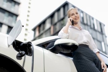 Focus charging-electric car with EV charger at charging station with blur businesswoman talking on phone with residential building in background as progressive lifestyle concept.. Focus electric car at charging station with blur background of progressive woman
