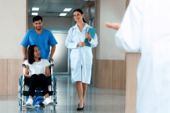 Doctor and male nurse transport a female patient in a wheelchair along sterile hospital corridor. Health care and nursing care for disabled handicapped patient in the hospital concept.. Doctor and nurse transport female patient in wheelchair at sterile corridor.
