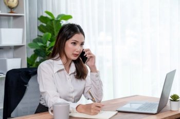 Young enthusiastic businesswoman talking on business call and writing notes on her laptop as multitask office lady. Female employee writing business tasks while talking on phone call with clients.. Young enthusiastic businesswoman talking on business call.