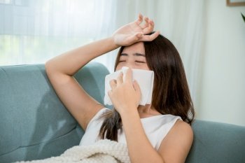 Sick woman sitting under blanket on sofa and sneeze with tissue paper in living room, Asian young female blowing nose sneezing in tissue at home, fever caught cold