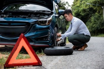 Asian businessman car broken has problems with wheel of his car during go to work in morning he changing replace rubber tire on wheels, business man have warning sign on road, problem transportation
