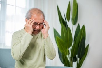 Headache. Sad Asian senior man sitting on sofa feeling hurt and lonely, elderly holds head with hand suffering from migraine headache, Old age health problems, healthcare