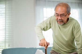Asian Old man with eyeglasses typing to stand up from sofa with walking cane, Elderly suffering from knee pain ache holding handle of cane, senior disabled man holding walking stick at home