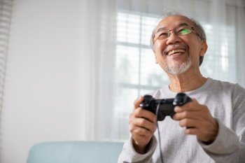 Asian senior old man enjoying holding joystick playing video game at home in living room, mature man hands using game controller, Funny on retirement elderly smile sitting on sofa life gaming