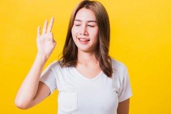 Asian Thai happy portrait beautiful cute young woman standing wear t-shirt showing gesturing ok sign with fingers looking to side, isolated studio shot on yellow background with copy space