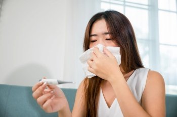 Sick Asian young woman sit under blanket on sofa she sneeze with digital thermometer and tissue paper at home, Cold flu and migraine concept, medical healthcare