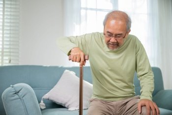 Asian senior old man with eyeglasses type to standing up from sofa with walking cane stick to walk at home, Elderly suffering from knee pain ache holding handle of cane, retirement medical healthcare