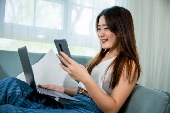 Happy woman use notebook browsing website and payment shopping online with smartphone, Asian young female using smart mobile phone while sitting on sofa in living room at home with laptop computer