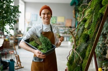 Portrait of young hipster florist smiling to camera standing at creative floral composition. Happy man decorator making decoration for home interior design. Portrait of young hipster florist standing at creative floral composition