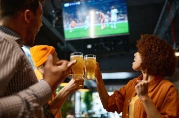 Happy friends drinking beer and cheering together in sports bar. Two young men and woman watching match and shouting, clinking glasses for win of favorite team on championship tournament. Happy friends drinking beer and cheering together in sports bar