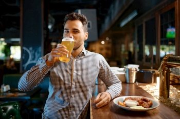 Satisfied man drinking cold fresh craft delicious beer while rest at sports bar. Young man standing at counter desk looking at camera while tasting foamy beverage. Satisfied man drinking cold fresh craft beer while rest at sports bar