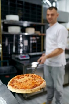 Young male chef holding freshly baked pizza on long shovel. Selective focus of hot Italian fast food. Young male chef holding freshly baked pizza on long shovel selective focus
