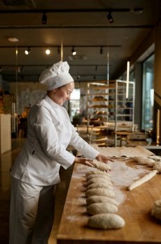 Young happy smiling woman baker in uniform forming bread loaves from raw dough at professional bakery kitchen. Woman baker forming bread loaves from raw dough at professional kitchen