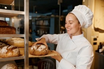 Beautiful woman baker in uniform checking freshly baked bread on tray rack at modern bakery kitchen. Beautiful woman baker in uniform checking freshly baked bread