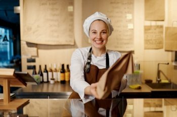 Portrait of friendly smiling bakery seller giving eco package with pastry standing at counter desk and looking at camera. Bakery shop concept. Portrait of friendly smiling bakery seller giving eco package with pastry