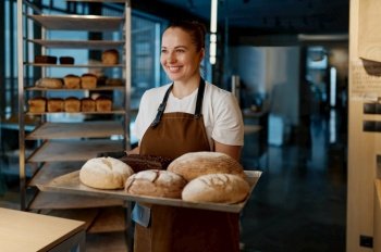 Pretty woman baker holding variety of baked bread on tray standing over bakery house background. Pretty woman baker holding variety of baked bread on tray