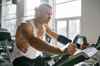 Healthy smiling sportsman using elliptical machine at gym fitness center. Happy motivated male bodybuilder doing aerobic exercise for slim and firm healthy body. Closeup view. Healthy smiling sportsman using elliptical machine at gym fitness center