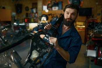 Portrait of young bearded repairman in coveralls carrying spare part of motorcycle frame on shoulder and looking at camera. Motorcycle garage repair service concept. Portrait of young repairman in coveralls carrying spare part of motorcycle