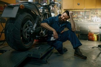 Satisfied smiling professional mechanic feeling glad looking at repaired motorcycle after good work in repair workshop. Satisfied smiling mechanic feeling glad looking at repaired motorcycle