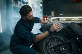 Young man worker wearing protective glasses sanding motorcycle from old paint while working at repair service. Young man repair service worker sanding motorcycle from old paint