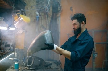 Serious concentrated repairman inspecting new gas tank welded for motorcycle. Serviceman working at smoky workshop in evening. Serious concentrated repairman inspecting new welded part for motorcycle