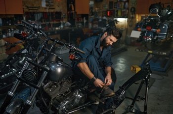 Background image on motorcycle workshop with auto mechanic resting during break time. Technician wearing overalls tying shoelaces on boots while sitting on motorbike. Background of motorcycle workshop with auto mechanic resting during break time