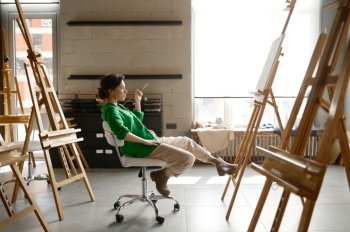 Female artist with paintbrush in hand looking seriously on her own picture while sitting on chair in art studio. Female artist with paintbrush in hand looking seriously on her own picture