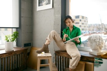 Happy smiling attractive woman sitting on windowsill looking aside holding paint palette in hands of her own workshop. Young female artist enjoying art leisure activity and creative hobby. Happy smiling woman sitting on windowsill holding paint palette in hands