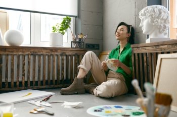 Relaxed young woman artist sitting on floor front of drawing accessories taking break and rest with eyes closed. Creative hobby enjoyment and searching inspiration concept. Relaxed young woman artist sitting on floor front of drawing accessories