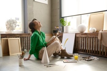 Young excited female artist laughing loudly while sitting on floor at home art studio. Attractive woman enjoying remote studying at online painter class hearing funny crazy joke. Young female artist laughing loudly sitting on floor at home art studio