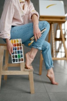 Cropped shot of barefoot female artist holding drawing tools, watercolor paints palette while sitting on chair at home art workspace. Cropped shot of barefoot female artist holding drawing tools sitting on chair