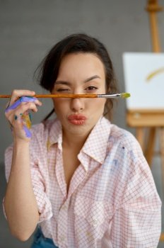 Portrait of funny woman artist holding paintbrush on cheerful and happy face winking looking at camera with positive expression. Portrait of funny woman artist holding paintbrush on face