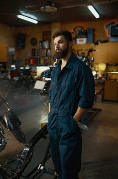 Motorcycle garage for repair and maintenance concept. Portrait of handsome serious repairman wearing overalls standing over workshop background. Portrait of handsome serious repairman wearing overalls standing in workshop